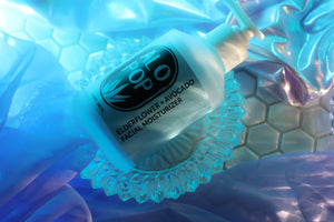 Elderflower and avocado facial moisturizer is laying on a glass dish white a blue light behind it. ALOPOP logo and product title is black text on a blue label.