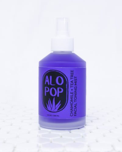 Chamomile + Tea Tree Toning Mist in a glass mist bottle. Liquid is purple and the label has the ALOPOP logo and title of this gentle face toning mist in black. The bottle is sitting on white tile with a white background.