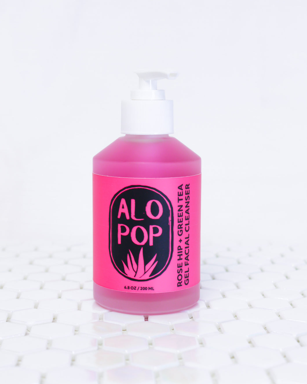 Glass bottle of Rose Hip and Green Tea Gel facial cleanser. Gel is hot pink with a white pump top lid and a hot pink label with black text on it. The text has the ALOPOP logo and product title. The gentle face wash bottle is sitting on geometric white tiles with the white background behind it.