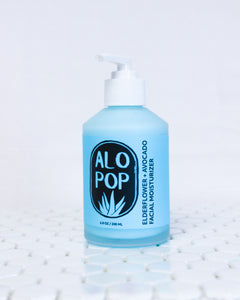 Glass bottle of Elderflower and Avocado Facial Moisturizer, creamy cyan blue lotion with a white pump top lid. Blue label with black ALOPOP logo and product title.  Gentle moisturizer is sitting on white geometric tiles with a white background.