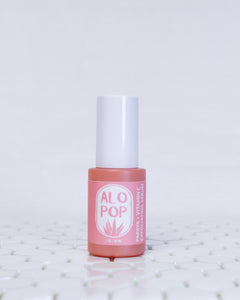 Papaya + Vitamin C Exfoliating Serum in a glass coral colored bottle with a white cap. The label of this glowing facial serum is pinky salmon colored with white text. The text has the ALOPOP logo and the product title. The serum bottle is sitting on geometric white tiles with a white background behind it. 