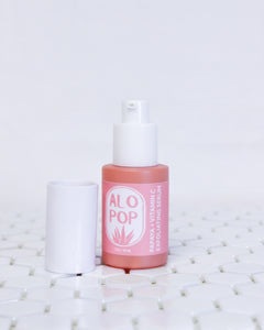 Papaya + Vitamin C Exfoliating Serum in a glass coral colored bottle with a white cap off sitting next to the bottle, the pump top of the bottle is also white. The label of this glowing facial serum is pinky salmon colored with white text. The text has the ALOPOP logo and the product title. The serum bottle is sitting on geometric white tiles with a white background behind it. 