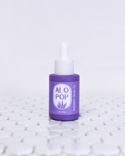 Glass Radiance Facial Oil bottle is sitting on white geometric tiles with a white background. The facial oil is in a muted lavender glass bottle with a matte finish. It has a white dropper top and a lavender label with white text on it. The text has the product title and the ALOPOP logo.