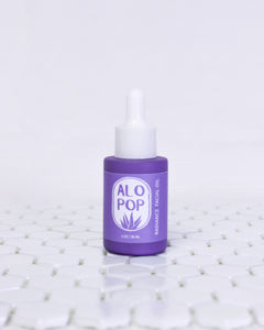 Glass Radiance Facial Oil bottle is sitting on white geometric tiles with a white background. The facial oil is in a muted lavender glass bottle with a matte finish. It has a white dropper top and a lavender label with white text on it. The text has the product title and the ALOPOP logo.