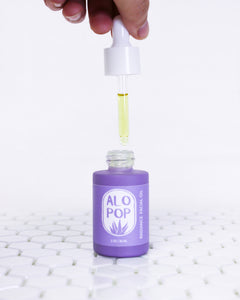 Glass Radiance Facial Oil bottle is sitting on white geometric tiles with a white background. The facial oil is in a muted lavender glass bottle with a matte finish. It has a white dropper top being help open with a person's hand. The dropper is full of the light yellow golden facial oil in a glass tube. It has a lavender label with white text on it. The text has the product title and the ALOPOP logo.
