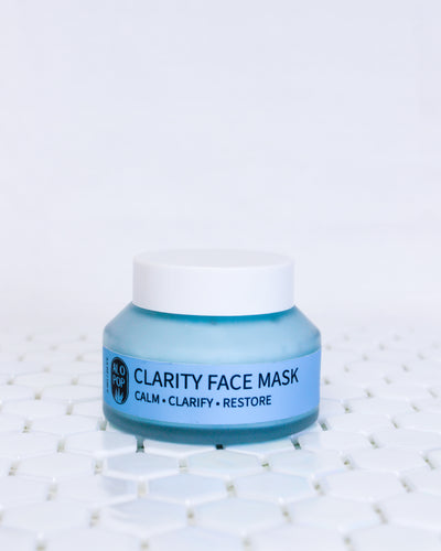 Blue clarity clay face mask in a glass jar with a white screw top lid. This gentle face mask is sitting on white geometric tiles with a white background behind it. The acne friendly mask has a blue label with black text.
