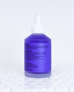 Chamomile + Tea Tree Toning Mist in a glass mist bottle. Liquid is purple and the label has the ingredients list, directions to use and address in black. The bottle is sitting on white tile with a white background.