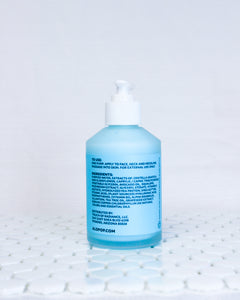Glass bottle of Elderflower and Avocado Facial Moisturizer, creamy cyan blue lotion with a white pump top lid. Blue label with black text about usage, ingredients list and ALOPOP address. Acne friendly moisturizer is sitting on white geometric tiles with a white background.