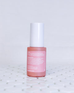 Papaya + Vitamin C Exfoliating Serum in a glass coral colored bottle with a white cap. The bottle is facing backwards so that you can see the label of this glowing facial serum that is pinky salmon colored with white text. The text has the usage instructions, ingredients and ALOPOP address/website. The serum bottle is sitting on geometric white tiles with a white background behind it. 