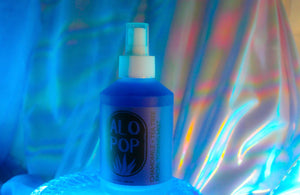 Chamomile + Tea Tree Toning Mist in a glass mist bottle. Liquid is purple and the label has the ALOPOP logo and title of this gentle face toning mist in black. The bottle is sitting on a glass dish with a dreamy rainbow haze behind it.