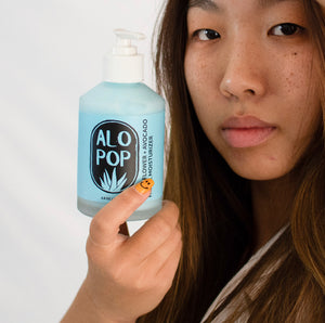 Person with long hair holding Elderflower and avocado Facial moisturizer in glass bottle up. finger nails are yellow with smiley faces, and they are looking at the camera. Gentle face lotion is blue.